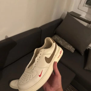 Nike Air Force 1 Low beige/brun - Nike Air Force 1 Low sneakers i beige med bruna swoosh-detaljer och röd minilogga på sidan. Klassisk perforerad tå, platt sula och AIR-text i rött på mellansulan. Skorna är tillverkade i skinn och har rund tå. Perfekt för dig som gillar stilrena och unika färgkombinationer.