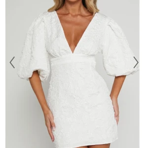 Vit kort klänning med puffärmar - Säljer min supersnygga studentklänning som är en vit kort klänning med djup v-ringning och bred midja. Klänningen har puffiga, korta ärmar och är tillverkad i ett strukturerat tyg som ger en lyxig känsla. Perfekt för dig som vill sticka ut med en trendig och feminin look.