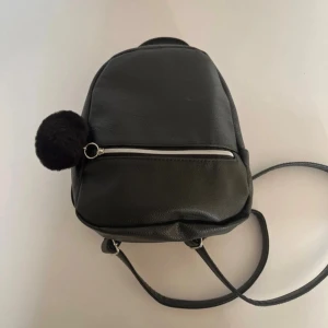 Svart ryggsäck med fluffig detalj - Snygg svart ryggsäck i skinnimitation med justerbara axelremmar och ett praktiskt handtag upptill. Väskan har en silverfärgad dragkedja framtill och en fluffig svart boll som dekoration. Perfekt storlek för att få med allt viktigt och stilren look.