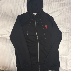 Svart hoodiejacka från Ami Paris - Svart hoodiejacka från Ami Paris med dragkedja, snörning i huvan och kängurufickor. På bröstet finns ett rött broderat hjärta med bokstaven A. Jackan är tillverkad i mjuk bomull och har långärmad passform. Storlek S