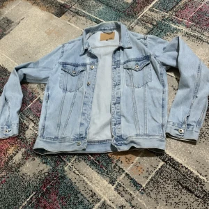 Ljusblå jeansjacka från 157 - Snygg ljusblå jeansjacka från 157 med klassisk krage, två bröstfickor med knappar och silverfärgade detaljer. Jackan har raka linjer och lång ärm, perfekt för dig som gillar en chill och tidlös stil. Passar till allt och är ett måste i garderoben.