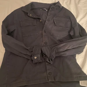 Mörkblå overshirt från Jack & Jones - Mörkblå  overshirt från Jack & Jones med två stora bröstfickor och knappar framtill. Skjortan har klassisk krage och långa ärmar med knapp vid manschetten. Pris kan diskuteras