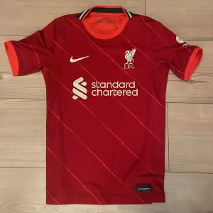 Liverpool FC röd matchtröja Nike - Liverpool FCs officiella matchtröja från Nike i rött med tunna diagonala ränder. Tröjan har korta ärmar, rund hals med orange detaljer och klubbmärke samt sponsortryck på bröstet. Tillverkad i lätt och ventilerande polyester för maximal komfort.