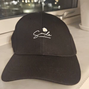 Svart keps med Smile-brodyr och blomma - Svart keps med böjd skärm och justerbart spänne bak. Framtill finns broderad text 'Smile' och en liten vit blomma ovanför. Kepsen är tillverkad i bomull och har en stilren, klassisk look som passar till många outfits.