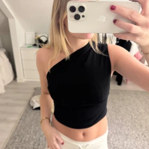 Svart oneshoulder croppad topp - Säljer en svart croppad topp med oneshoulder-design. Toppens stil är riktigt snygg och trendig, med en asymmetrisk axel som ger en cool vibe. Materialet känns mjukt och stretchigt, perfekt för att skapa en clean look. Passar dig som gillar att sticka ut med enkla detaljer.