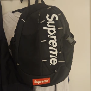 svart ryggsäck med logga -  Ryggsäck i svart med stor vit logga längs framsidan och klassisk röd label längst ner. Ryggsäcken har flera fack, justerbara axelremmar och detaljer i syntetmaterial. Perfekt för dig som vill ha streetstyle och funktion i ett.