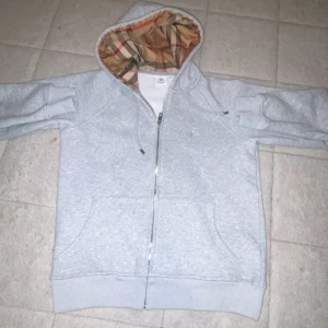 Grå Burberry hoodie med rutig huva - Säljer en grå hoodie från Burberry med dragkedja och klassisk rutig huva i beige, svart och rött. Tröjan har broderad Burberry-logga på bröstet, snörning i huvan och två stora fickor framtill. Perfekt för dig som vill ha en stilren och lyxig look.