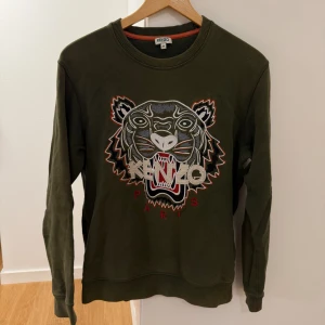 Grön Kenzo sweatshirt med tiger - Säljer en grön sweatshirt från Kenzo i storlek M med det ikoniska broderade tigermotivet och Kenzo Paris-text på bröstet. Tröjan har rund hals, ribbade muddar och är långärmad. Perfekt för dig som gillar streetwear och vill sticka ut med snygg design.