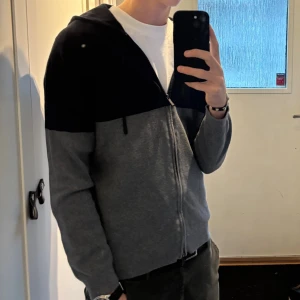 Grå och svart hoodie från Massimo Dutti - Snygg tvåfärgad hoodie från Massimo Dutti i grått och svart. Tröjan har dragkedja framtill, huva med snörning och är tillverkad i en mjuk mix av bomull och silke. Perfekt för chill dagar eller när du vill ha en stilren look. 