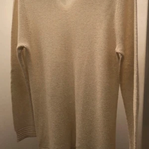 Beige stickad tröja med v-ringning - Snygg beige stickad tröja från Marina med v-ringad hals och långärmade ärmar. Tröjan har ett diskret ribbat mönster och är gjord i ett mjukt material som känns skönt mot huden. Perfekt för dig som gillar enkel och stilren design.