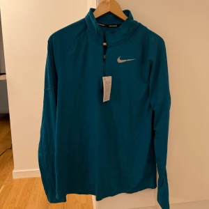 Blå Nike Dri-Fit långärmad träningströja - Nike Dri-Fit långärmad träningströja i blått med halv dragkedja framtill och reflekterande logga på bröstet. Perfekt för löpning och sport, tillverkad i lätt polyester som andas. Snygg och funktionell design med meshpaneler för extra ventilation.