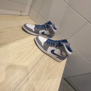 Nike Jordan 1 Mid blå/grå sneakers - Nike Jordan 1 Mid sneakers med vit och grå ovandel i skinn, blå detaljer och snörning. Swoosh-logga i blått på sidan, blå yttersula och klassisk Air Jordan Wings-logga på ankeln. Perfekt för dig som gillar streetwear och vill sticka ut med snygga färger.