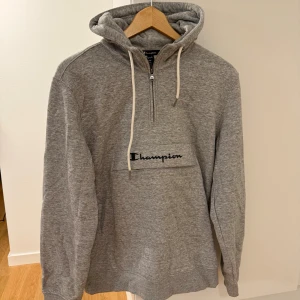 Grå hoodie från Champion med half zip - Grå hoodie från Champion i storlek medium. Tröjan har huva med snörning, half zip framtill och broderad Champion-logga på bröstet. Tillverkad i mjuk bomullsmix och har långa ärmar. Perfekt för chill dagar och streetwear-stil.