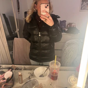 Svart Moncler pufferjacka med huva - Säljer min svarta pufferjacka från Moncler med huva och dragkedja framtill. Jackan har en glansig finish, Moncler-logga på ärmen och är perfekt när det är kallt ute. Passformen är normal och jackan har långa ärmar. Snygg och stilren look! Storlek 14y