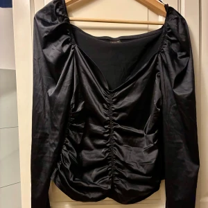 Svart omlottopp från Gina Tricot - Snygg svart omlottopp från Gina Tricot med lång ärm och v-ringad hals. Toppen har rynkade detaljer framtill och är tillverkad i mjuk polyester med lite stretch. Perfekt för dig som gillar trendiga och stilrena plagg med en twist.