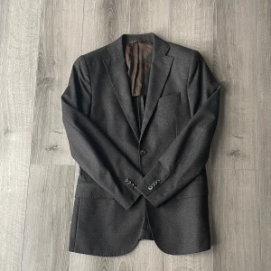 Mörkbrun kavaj i Loro Piana dreamtweed  - Snygg mörkbrun kavaj sydd av j.lindeberg med klassisk passform. Tillverkad i 100% extrafin ull från Loro Piana, med två framfickor och en två knappstängning. Perfekt för dig som vill ha en stilren och tidlös look.