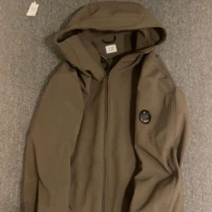 Brun vindjacka från C.P. Company - Säljer en brun vindjacka från C.P. Company med huva och dragkedja framtill. Jackan har den klassiska linsen på ärmen och en stilren design. Materialet är syntet och jackan har en normal passform med långärmade ärmar. Perfekt för dig som gillar streetwear och snygga detaljer. Den är storlek S men passar som M. Den har gått sönder som ni ser på bild 3 .
