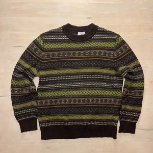 Mönstrad brun stickad tröja H&M - Säljer en brun stickad tröja från H&M med gröna, gula och grå detaljer i ett klassiskt mönster. Tröjan har rund halsringning, långa ärmar och ribbade muddar. Perfekt för dig som gillar retrovibbar och vill ha något unikt i garderoben.