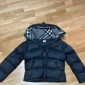 Svart pufferjacka från Burberry - Säljer en svart pufferjacka från Burberry med avtagbara ärmar och huva med klassiskt rutigt foder i grått, vitt och rött. Jackan har dragkedja framtill, två sidofickor med dragkedja och en snygg Burberry London England-patch på ärmen. Perfekt för kalla dagar.