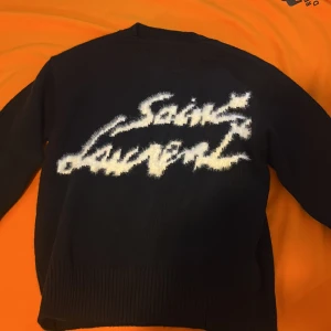 Svart Saint Laurent stickad tröja - Cool svart stickad tröja från Saint Laurent med vit, fluffig logga framtill. Tröjan har rund hals, långa ärmar och ribbade muddar vid ärmslut och nederkant. Perfekt för dig som vill sticka ut med en statement piece.