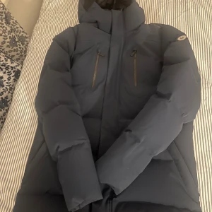 Mörkblå pufferjacka med huva från sail racing  - Säljer en mörkblå pufferjacka med huva och två vertikala bröstfickor med dragkedja. Jackan har en quiltad design med breda paneler och är långärmad. Perfekt för kalla dagar och har en clean, modern look. Jackan går för cirka 6000 kr ny jag säljer den för mindre  än halva priset i inprincip ny skick 