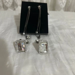 Mix av örhängen och halsband med kristaller - Fyra olika smycken: ett par fyrkantiga örhängen med stora klara stenar och små gnistrande stenar runt, ett par långa silverfärgade örhängen med pärlor, ett halsband med en glittrig ormberlock och ett guldigt halsband med bladformade kristaller. Perfekt för dig som gillar bling och unika detaljer.