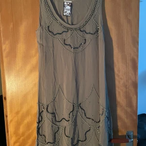 Grön ärmlös klänning med pärlor - Snygg grön ärmlös klänning från Vintage Stories med rund halsringning och dekorativa pärlor och broderade detaljer framtill. Klänningen har ett unikt mönster med pärlor i vågformade linjer som ger en cool vintagekänsla. Perfekt för dig som gillar statement-plagg.