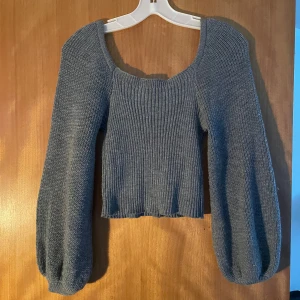 Grå stickad topp med puffärmar - Säljer en grå stickad topp med bred rund halsringning och puffiga långärmar. Toppen har ribbad struktur framtill och är croppad i modellen, vilket ger en trendig och cool vibe. Perfekt för dig som gillar att sticka ut med snygga detaljer.