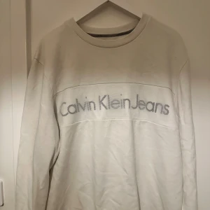 Vit sweatshirt från Calvin Klein Jeans XL - Vit sweatshirt från Calvin Klein Jeans i storlek XL. Tröjan har ett stort tryck med loggan över bröstet och ribbade muddar vid halsen. Mjuk och skön bomullskvalitet, perfekt för chill dagar. Enkel och stilren design som funkar till jeans eller joggers.