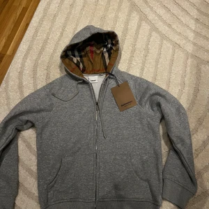 Grå hoodie från Burberry med dragkedja - Säljer en grå hoodie från Burberry med klassiskt rutigt foder i huvan och broderad logga på bröstet. Tröjan har dragkedja framtill, snörning i huvan och två fickor. Perfekt för dig som gillar stilrena och exklusiva plagg.