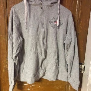 Grå hoodie Polo Club XL - Grå hoodie från Polo Club Saint Vincent i storlek XL. Tröjan har huva med snörning, dragkedja framtill och två fickor. Liten logga med amerikansk flagga på bröstet. Mjuk och skön bomullsmix, perfekt för chill dagar.