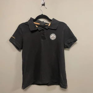 Svart Mercedes-Benz pikétröja - Svart pikétröja från Mercedes-Benz med broderad logga på bröstet och guldfärgad detalj på insidan av kragen. Kortärmad modell med klassisk krage och knappar framtill. Snyggt broderad 'Mercedes' på ärmen. Perfekt för dig som gillar bilar och stil.