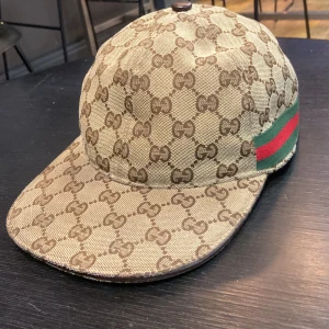 Gucci keps med GG-mönster, S - Snygg beige och brun keps från Gucci med klassiskt GG-monogram över hela kepsen. På sidan finns den ikoniska gröna och röda randen. Kepsen är tillverkad i bomull och polyester, har böjd skärm och en cool streetstil. Perfekt för dig som vill sticka ut med lyxig vibe. 
