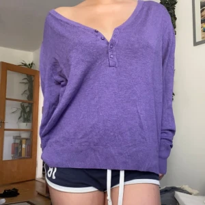 Lila långärmad tröja från B.Young - Lila långärmad tröja från B.Young med v-ringning och fyra knappar framtill. Tröjan har en loose passform och är tillverkad i mjuk bomullsmix. Perfekt för dig som gillar chill och avslappnad stil med lite djupare halsringning.