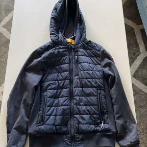 Blå quiltad jacka från Parajumpers - Snygg mörkblå quiltad jacka från Parajumpers med huva och dragkedja. Jackan har ribbade muddar, två sidofickor med dragkedja och en bröstficka. Ärmarna är i mjukare material och axeln pryds av Parajumpers-logga. Den här 2 små hål och en liten fläck som går bort om man försöker och pris ej hugget i sten 🪨 