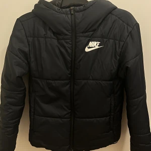 Svart pufferjacka från Nike XS - Svart pufferjacka från Nike i storlek XS. Jackan har huva, vita Nike-loggan på bröstet och dragkedja framtill. Quiltad design med långa ärmar och två sidofickor. Perfekt för kyliga dagar och har en clean, sportig look.