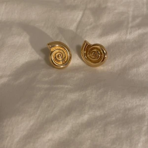 Guldiga spiralformade örhängen aldrig använd  - Säljer ett par coola örhängen i guldig metall med spiralformad design. De har en snygg, snäckinspirerad form som verkligen sticker ut och ger en unik touch till din stil. Perfekta för dig som gillar statement-smycken och vill ha något annorlunda.