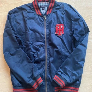 Mörkblå bomberjacka med röd patch - Snygg mörkblå bomberjacka från Tommy Hilfiger med röd och svart randig krage, muddar och nederkant. Stor röd TH-patch på bröstet, dragkedja framtill och två sidofickor. Perfekt för dig som gillar college-vibe och sportig stil.