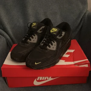 Nike Air Max 90 svart/svarta sneakers  -  Svarta Nike Air Max 90 sneakers med svarta ditsljer och neon-gula loggor. Klassisk chunky sula med synligt Air Max-fönster och platt yttersula. Ovandel i syntet och skinn, snörning framtill och rund tå. Perfekt för dig som gillar streetstyle och sportig vibe. Dom har lite mocka på sig som man kan fixa lite 