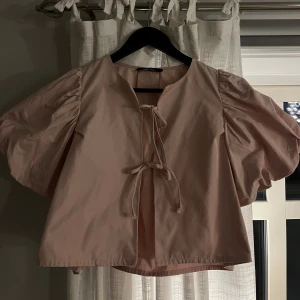 Ljusrosa blus med puffärm och knyt - Säljer en ljusrosa blus från Gina Tricot med puffiga korta ärmar och två knytband framtill. Blusen har en rak passform och är tillverkad i mjuk bomull. Perfekt för dig som gillar voluminösa ärmar och romantisk stil.