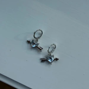 Silverörhängen med hjärta och pil - Ett par silverfärgade örhängen med hjärtformad glittrande sten och en pil som går igenom hjärtat. Perfekt accessoar för dig som gillar unika detaljer och romantisk stil.