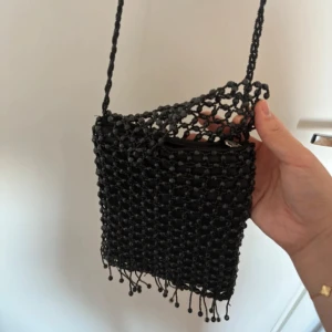 Vintage axelväska med pärldetaljer - Unik svart axelväska med handgjord känsla, täckt av små svarta pärlor och fransar längst ner. Väskan har ett innerfack med dragkedja och en flätad axelrem. Perfekt för dig som vill sticka ut med en cool accessoar.