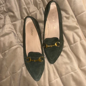 Mörkgröna mocka loafers från Gabor - Snygga loafers från Gabor i mörkgrön mocka med spetsig tå och guldfärgade metallspännen framtill. Använda fåtal gånger, säljer pga att dem är för små. 