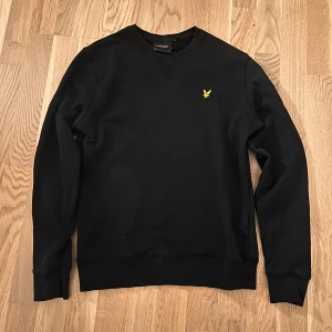 Svart sweatshirt från Lyle & Scott - Svart sweatshirt från Lyle & Scott med klassisk rund hals och gul broderad logga på bröstet. Tröjan har långa ärmar och ribbade muddar vid ärmslut och nederkant. Perfekt för en clean och stilren look. Storlek XS Fint Skick. Priset är inte hugget i sten!