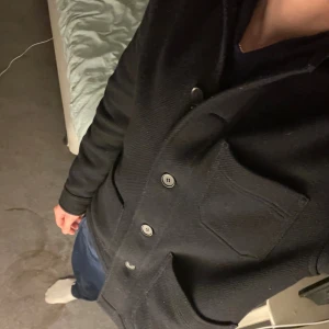 Svart overshirt med knappar - Svart overshirt med klassisk krage och tre framfickor. Jackan har en rak och loose passform, stängs med stora knappar framtill och är långärmad. Materialet ser ut att vara tjockt och lite strukturerat, perfekt för lager-på-lager under höst och vinter.