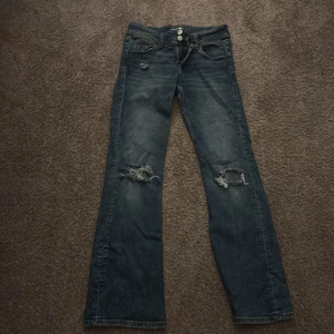 Blå bootcut jeans från Gina Tricot - Snygga blå jeans från Gina Tricot i bootcut-modell med slitna detaljer och hål vid knäna. Klassisk femficksdesign, dubbla knappar i midjan och coola fickor med knapp bak. Perfekt för dig som gillar en avslappnad och trendig look.