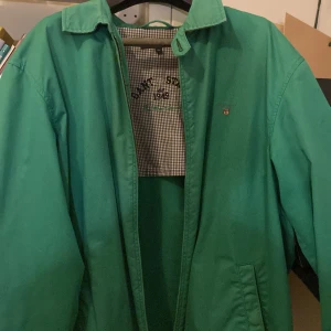 Grön vindjacka från GANT - Snygg grön vindjacka från GANT med klassisk krage och broderad logga på bröstet. Insidan har ett rutigt foder med broderad text 'GANT STATE 1949 The Windcheater'. Jackan har långa ärmar och praktiska fickor framtill. Perfekt för dig som gillar stilren och sportig look. Även lite vintege, vilket är populärt nu. Storleken är xl men passar även l och ibland m