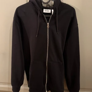 Svart zip hoodie  från Weekday - Svart hoodie från Weekday med dragkedja framtill, huva med snörning och två stora fickor. Klassisk modell i mjuk bomullsmix, perfekt för chill dagar. Långärmad och med ribbade muddar för extra komfort.