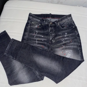 Svarta Dsquared2 jeans med färgstänk - Snygga svarta jeans från Dsquared2 med coola färgstänk i vitt och rött samt slitna detaljer. Klassisk femficksmodell med Dsquared2 patch bak i rött och gult. Jeansen har en rak passform och är tillverkade i jeansmaterial.