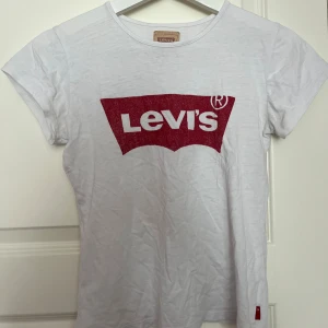 Vit Levi's t-shirt med röd logga - Snygg vit t-shirt från Levi's med den klassiska röda loggan tryckt framtill. Modellen har rund hals och korta ärmar, perfekt för en chill och stilren look. Materialet är mjuk bomull som känns skönt mot huden. Passar dig som gillar ikoniska streetwear-plagg.